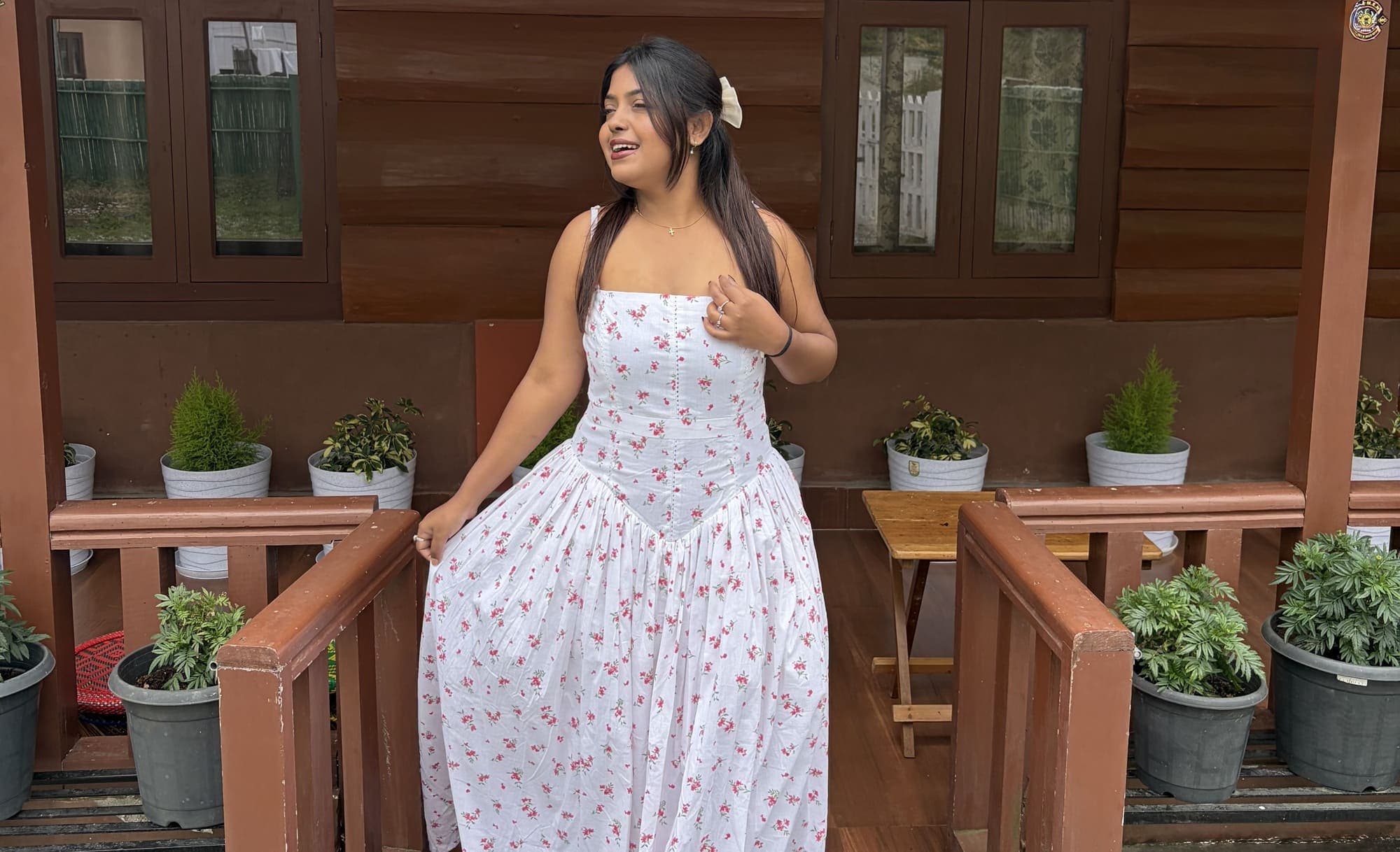A model in a white floral dress standing on a wooden veranda lined with potted plants