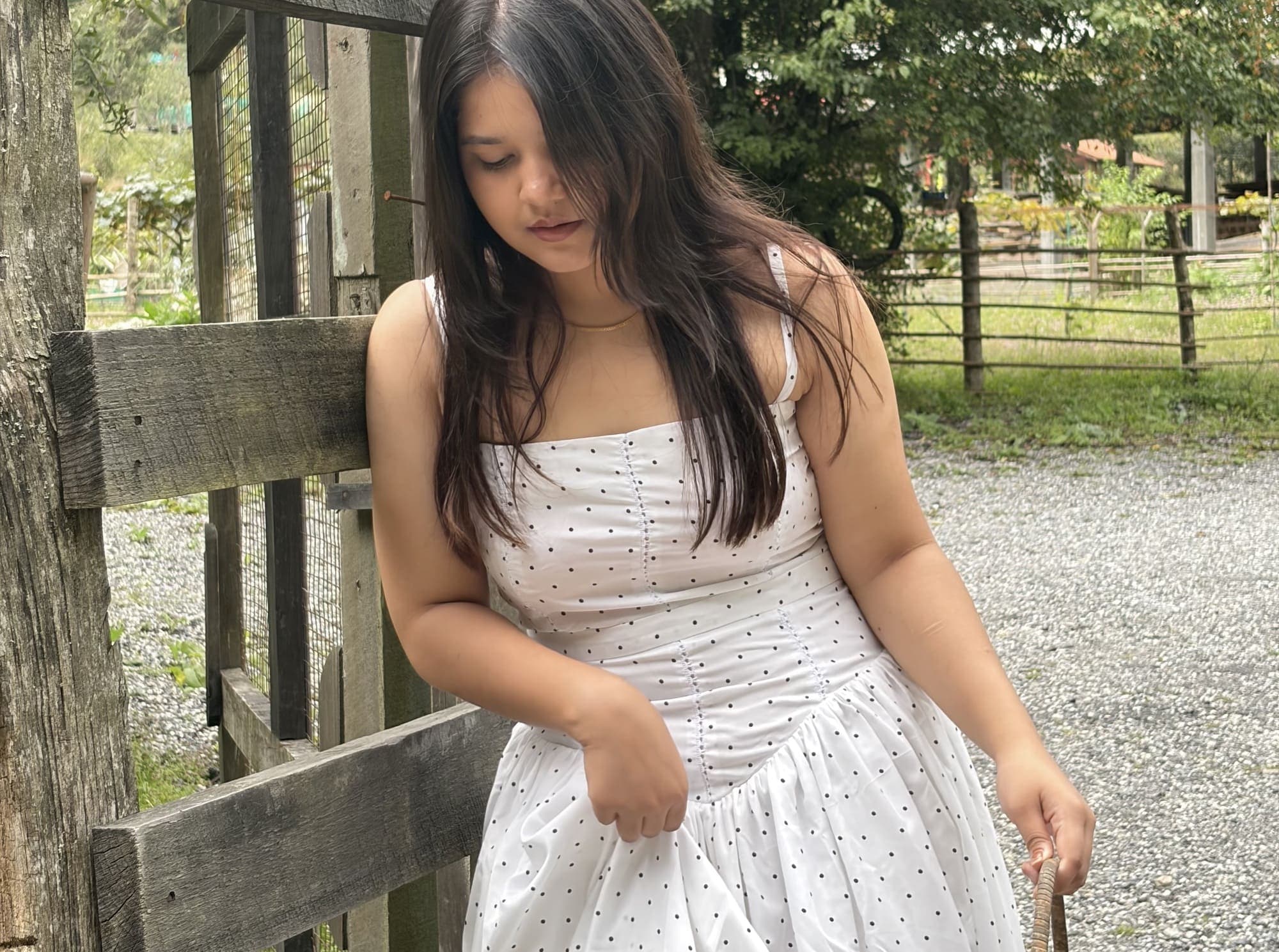 A model in a white polka-dot dress leaning against a weathered wooden fence in dappled sunlight