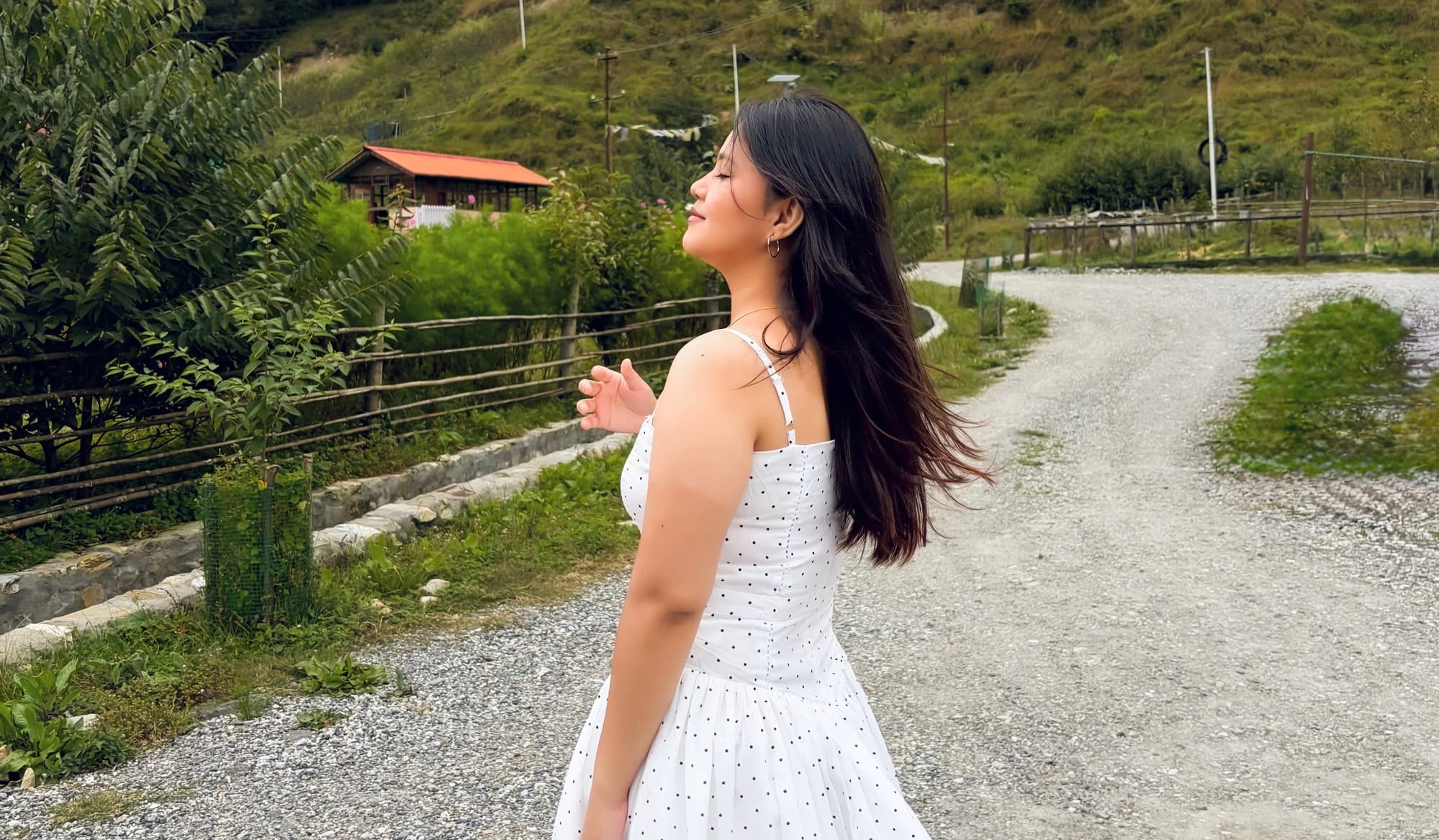 A model in a white polka-dot dress walking along a gravel garden path with a green hillside behind her